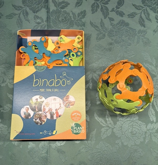 Geöffnete Binabo-Schachtel mit bunten Chips aus Bioplastik, daneben ein fertig gesteckter Ball in Orange und Grün auf einer Tischdecke
