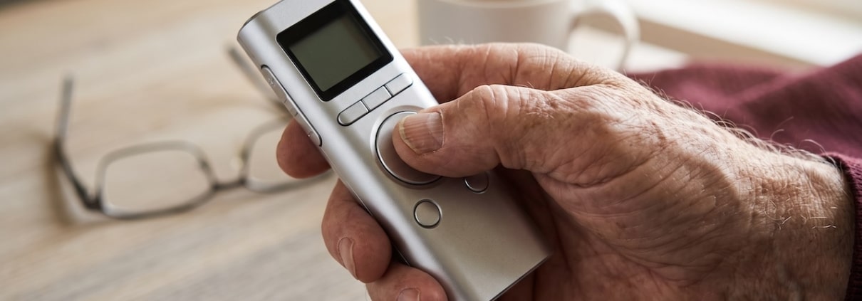Hand eines älteren Mannes hält ein Diktiergerät mit großen Tasten, der Daumen liegt auf der Aufnahmetaste
