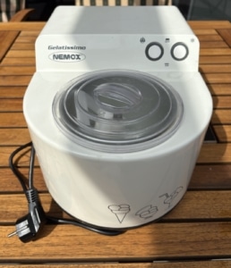 Weiße Eismaschine Nemox Gelatissimo mit zwei Drehknöpfen und durchsichtigem Deckel auf einem Holztisch im Garten