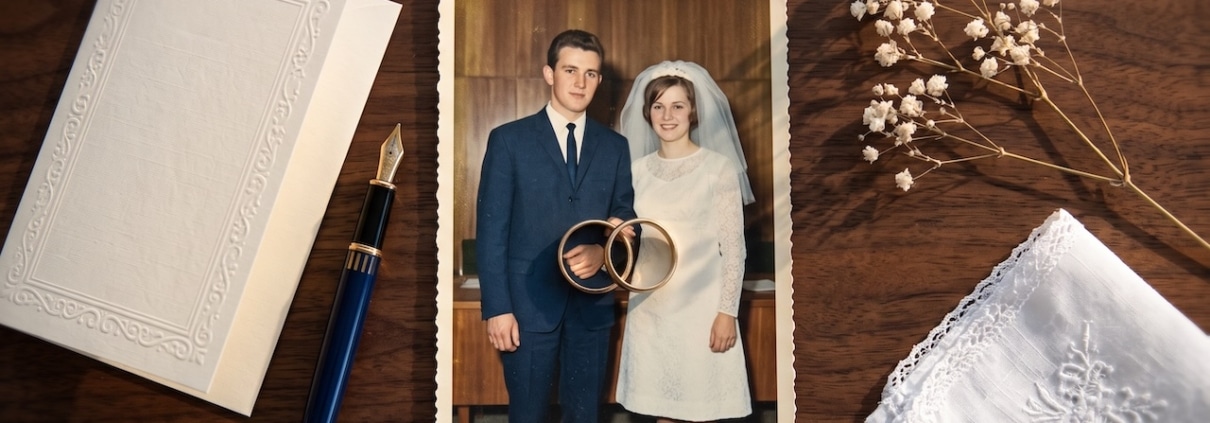 Zwei Eheringe auf einer Hochzeitsfotografie aus den sechziger Jahren, daneben eine Karte, ein Füllfederhalter und getrocknete Blüten