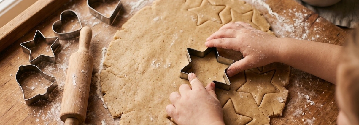 Kinderhände stechen Sterne aus ausgerolltem Plätzchenteig auf einer bemehlten Holzplatte, daneben liegen Ausstechformen und ein Nudelholz