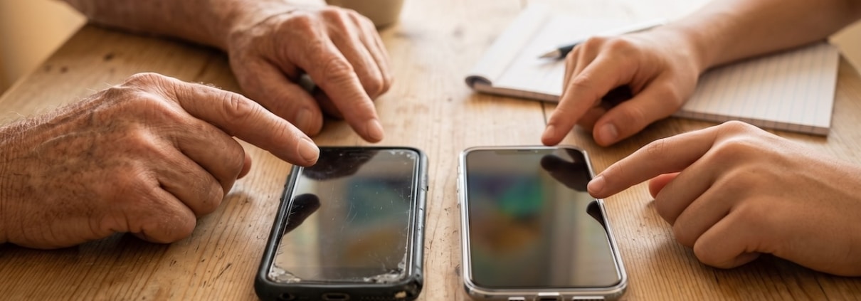 Ein altes und ein neues Smartphone liegen auf dem Tisch, die Hände von Großvater und Enkel deuten auf die Geräte