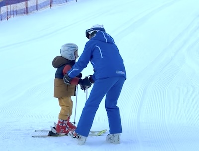 Ein kleines Kind in Skikleidung bei seiner ersten Skistunde, der Skilehrer in Blau hält es an den Händen – auf einer flachen Piste in Südtirol